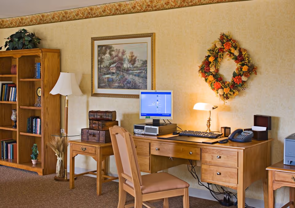 A cozy office corner with a wooden desk and chair. On the desk, there is a computer with a keyboard, a telephone, a small lamp, and a tissue box. Above the desk, a colorful floral wreath hangs on the wall. To the left, there is a wooden bookshelf filled with books and decorative items, and a floor lamp with a white shade. A framed painting of a countryside scene is mounted on the wall above a small side table with decorative boxes.
