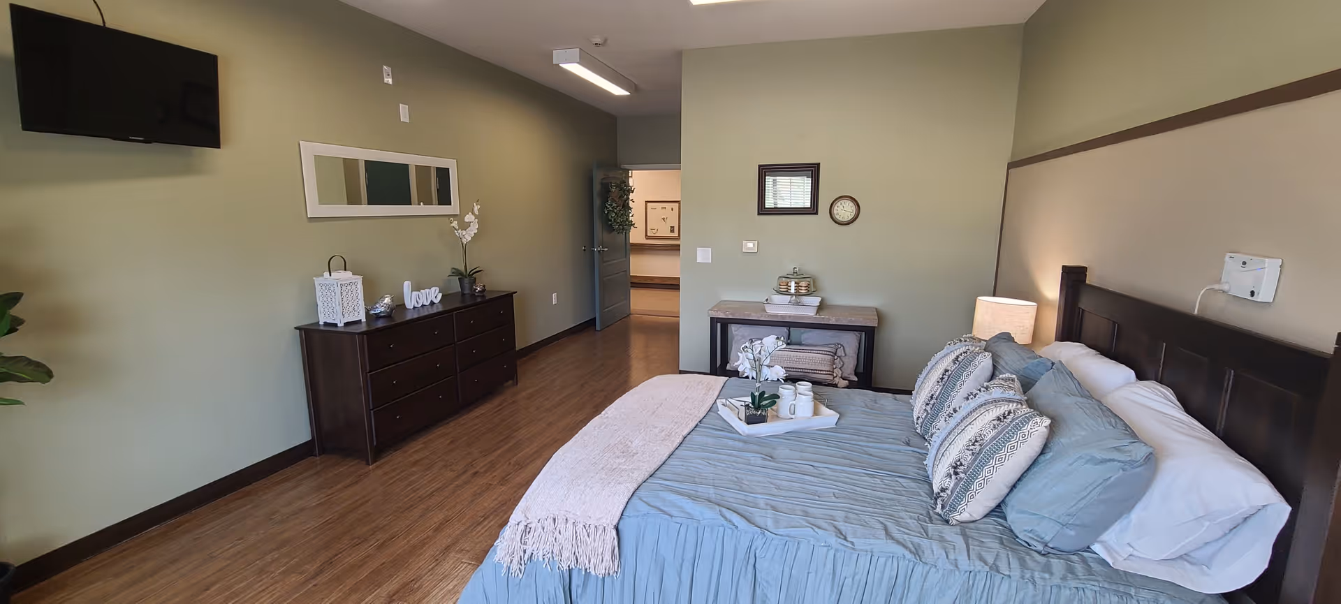 A neatly arranged bedroom with a large bed featuring blue bedding and decorative pillows. There is a wooden dresser with decorative items including a white lantern, a 'love' sign, and a small plant. A flat-screen TV is mounted on the green wall above the dresser. The room has wooden flooring and a small table with folded towels and a clock on the wall above it. The door to the room is open, showing a glimpse of another room.