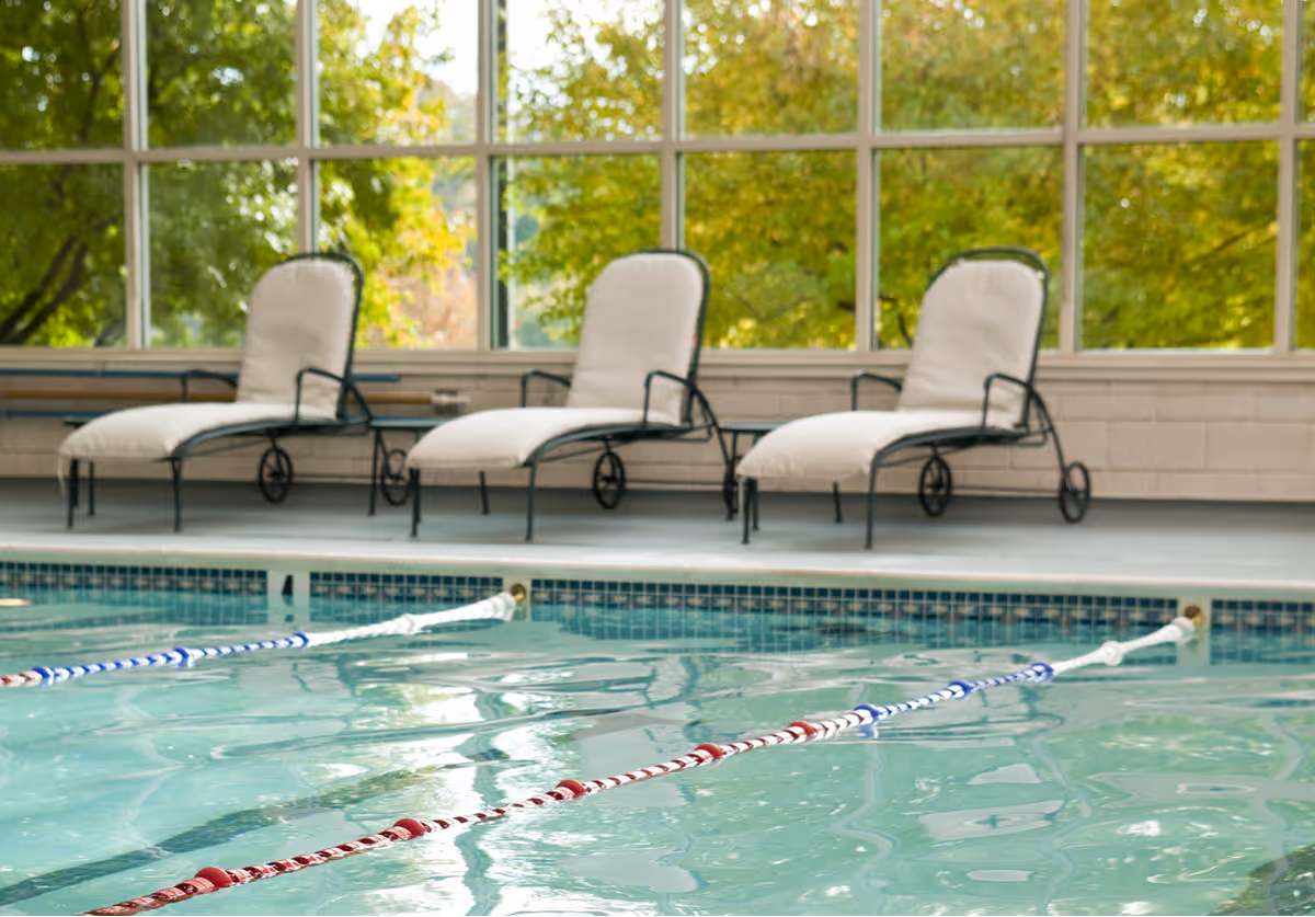 Indoor swimming pool with three cushioned lounge chairs lined up against a large window showing green trees outside.