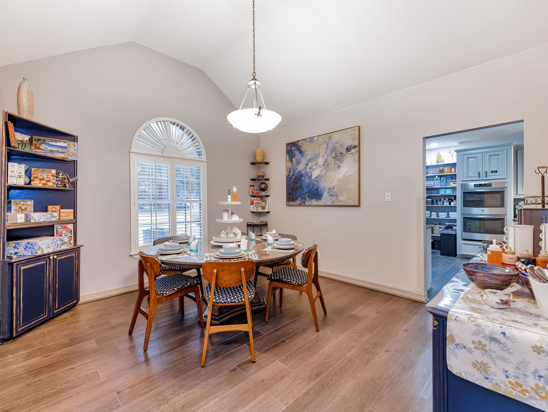 A dining room with a round table set for six people, featuring plates, glasses, and napkins. The room has wooden flooring, a large arched window with white shutters, a hanging light fixture, and a blue cabinet filled with books and decorative items. A doorway leads to a kitchen area with white cabinetry and stainless steel appliances.