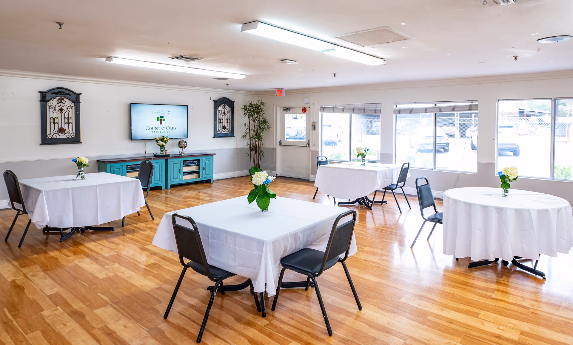 A bright dining room with wooden floors featuring several tables covered with white tablecloths and small floral centerpieces. Black chairs are arranged around the tables. A turquoise cabinet with a TV mounted above it is visible on the far wall, displaying the text 'Country Oaks Care Center'. Large windows let in natural light, and decorative wall hangings and a potted plant add to the room's decor.