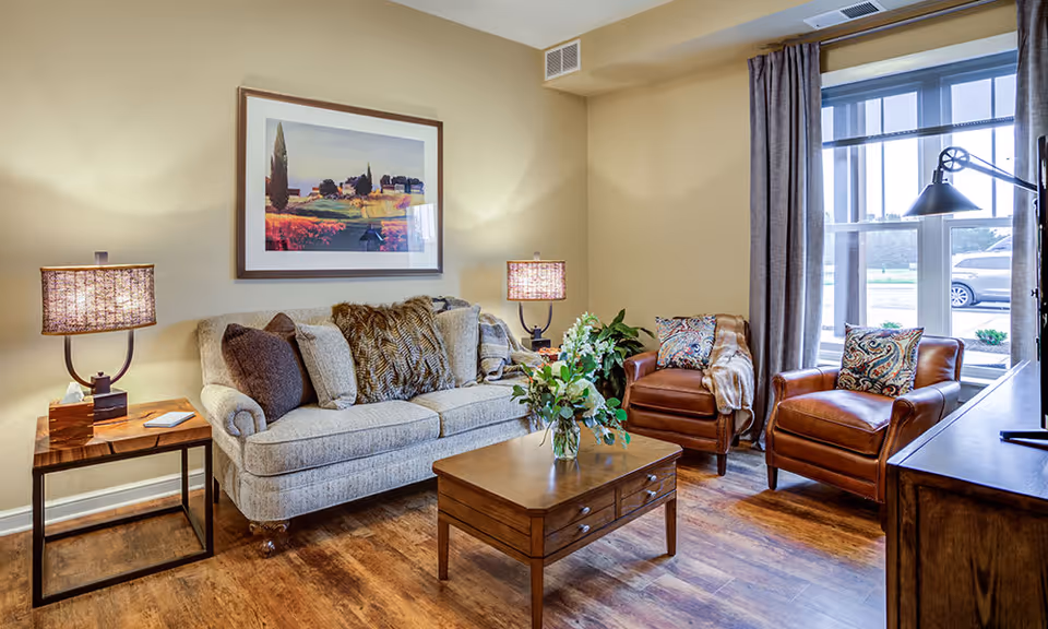 A cozy living room with a beige sofa adorned with multiple cushions, two brown leather armchairs with patterned pillows, a wooden coffee table with a vase of flowers, two side tables each with a lamp, a large window with gray curtains, and a framed landscape painting on the wall.