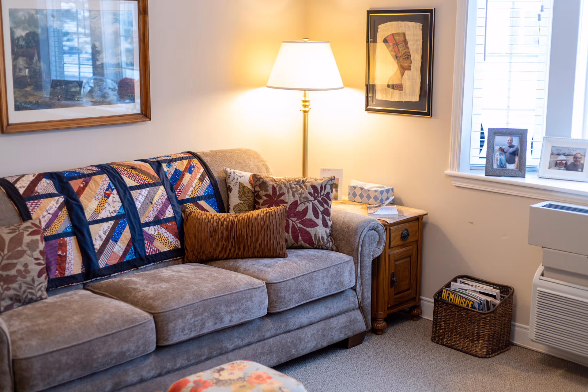 A cozy living room area featuring a beige sofa adorned with patterned cushions and a colorful quilt draped over the back. Next to the sofa is a wooden side table with a lamp, tissue box, and some papers. On the wall above the side table hangs framed artwork. A window with white trim lets in natural light, and a basket filled with magazines sits on the floor near a heating or air conditioning unit.