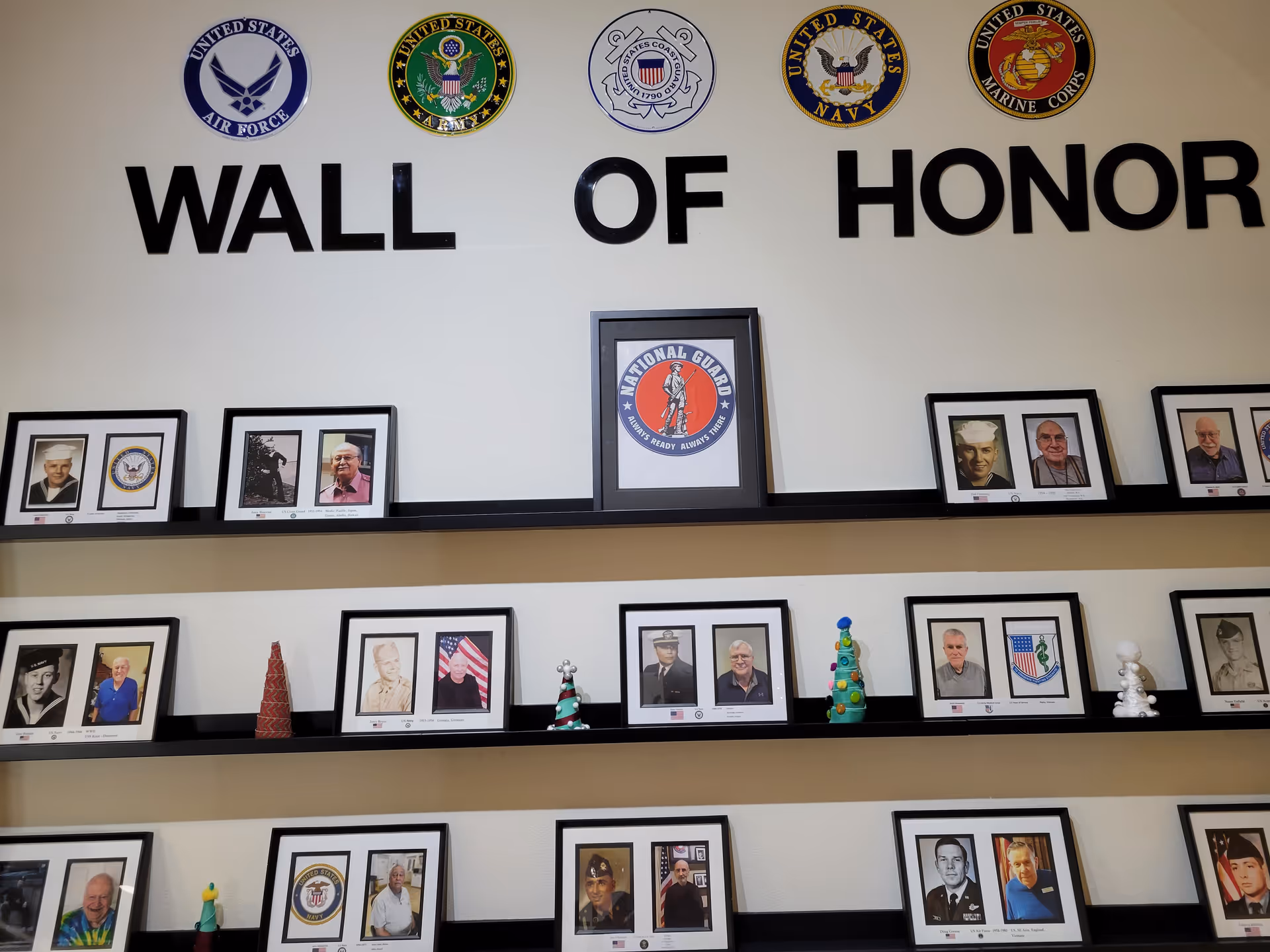 A Wall of Honor display featuring framed photographs of veterans alongside military branch emblems including the United States Air Force, Army, Coast Guard, Navy, Marine Corps, and National Guard. The photos are arranged on three black shelves mounted on a beige wall, with small decorative items placed between some frames.