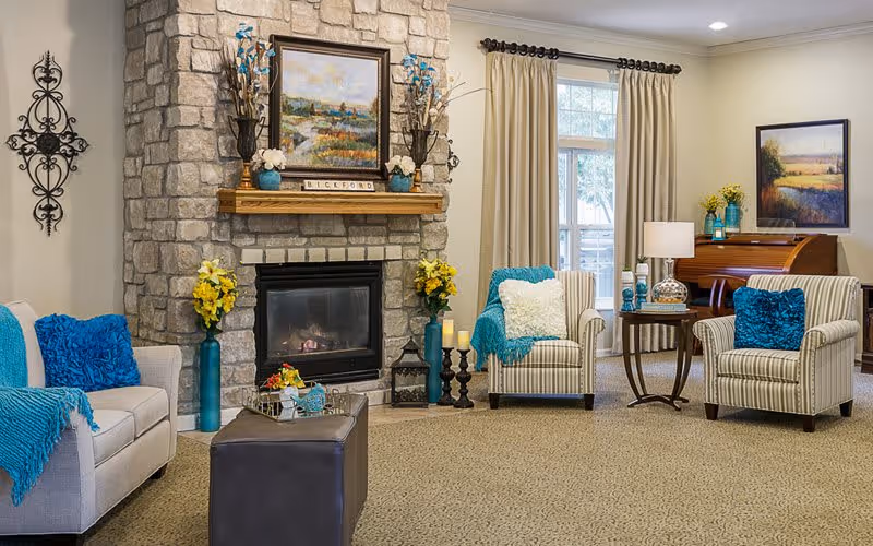 A cozy living room with a stone fireplace in the center, decorated with vases of flowers and a framed painting above the mantel. There are two striped armchairs with blue and white pillows and a beige sofa with blue pillows and a blue throw blanket. A small wooden table with a lamp and decorative items sits between the armchairs. A window with beige curtains lets in natural light, and a piano with a framed painting above it is visible in the background.
