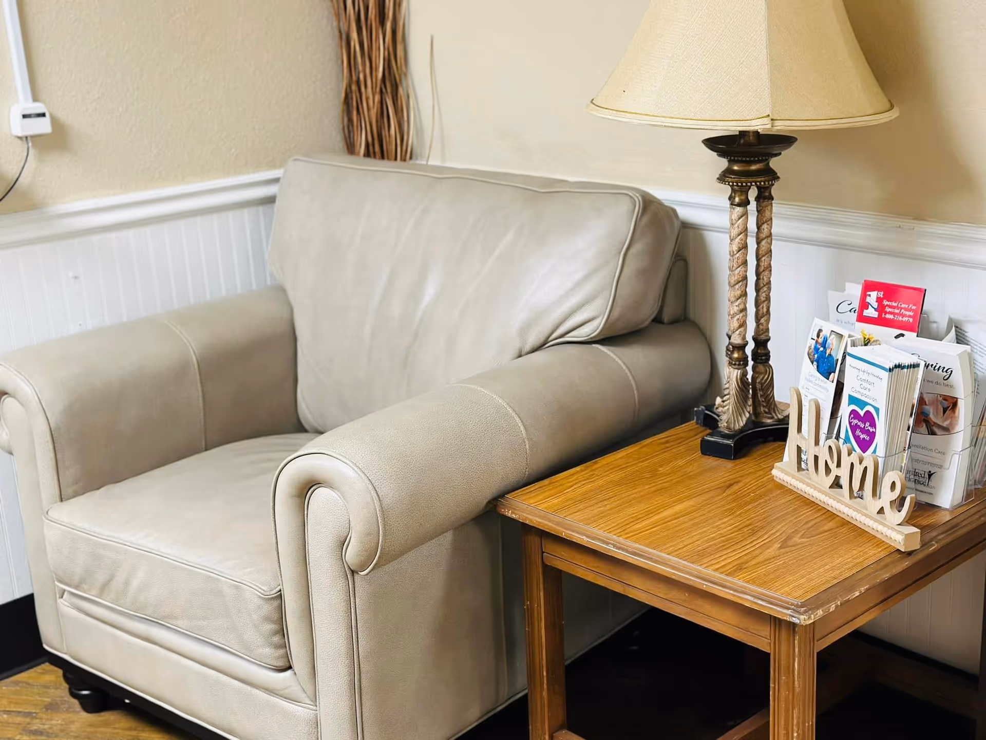 A beige leather armchair next to a wooden side table with a decorative lamp, a wooden 'Home' sign, and a brochure holder containing various pamphlets, set against a cream-colored wall with white wainscoting.