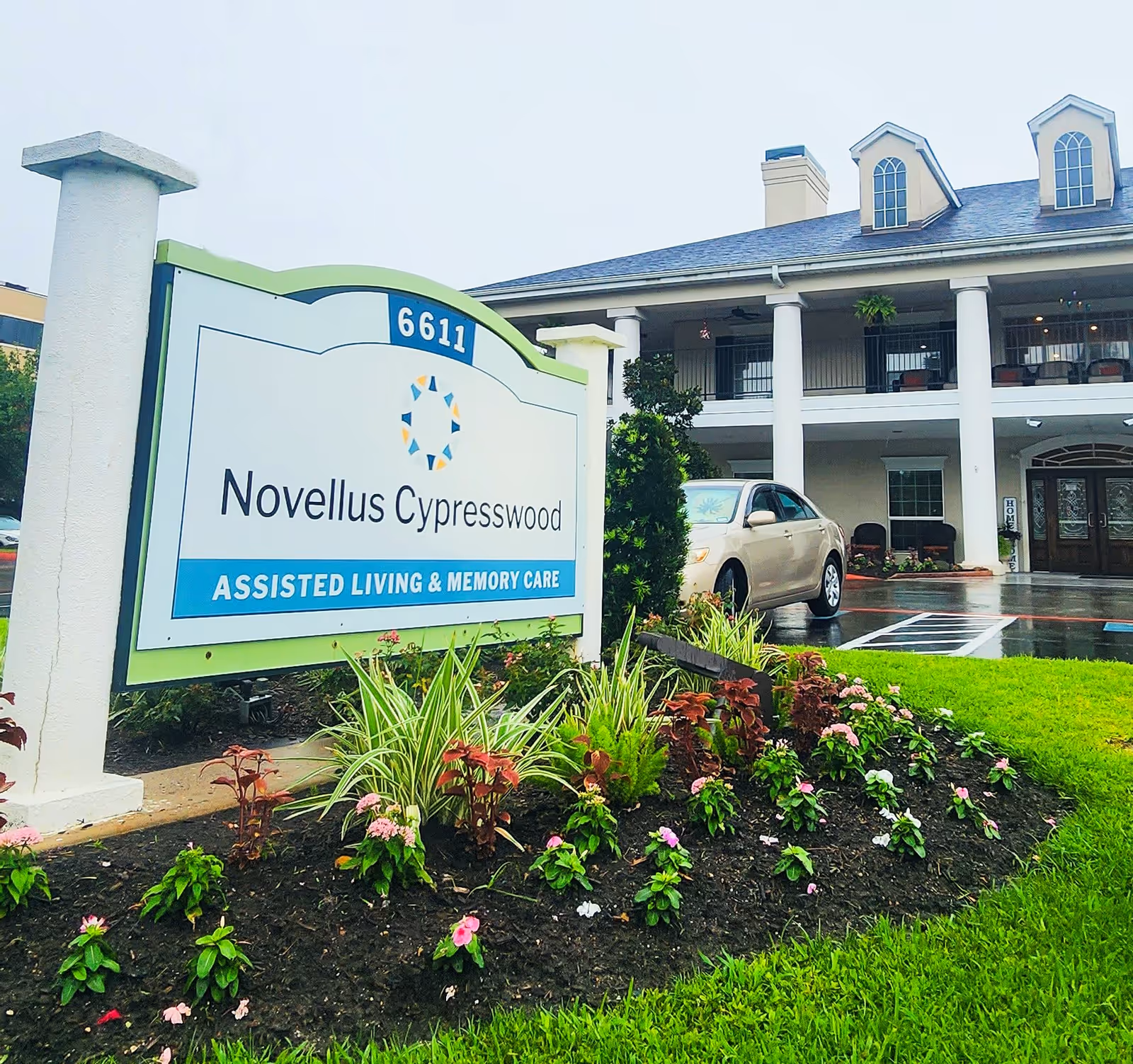 Front sign reading "Novellus Cypresswood Assisted Living & Memory Care" with a landscaped flowerbed and the facility building and entrance in the background.