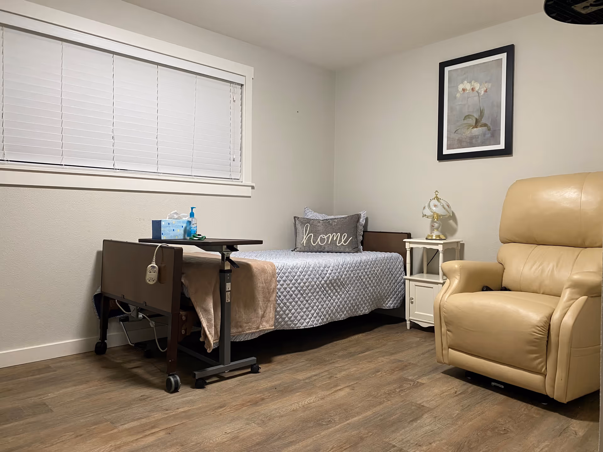 A small, cozy bedroom with a single bed covered in a gray quilt and a pillow with the word 'home' on it. Next to the bed is a white nightstand with a decorative lamp. A beige recliner chair is positioned to the right. The room has wooden flooring, a window with closed white blinds, and a framed floral picture on the wall.