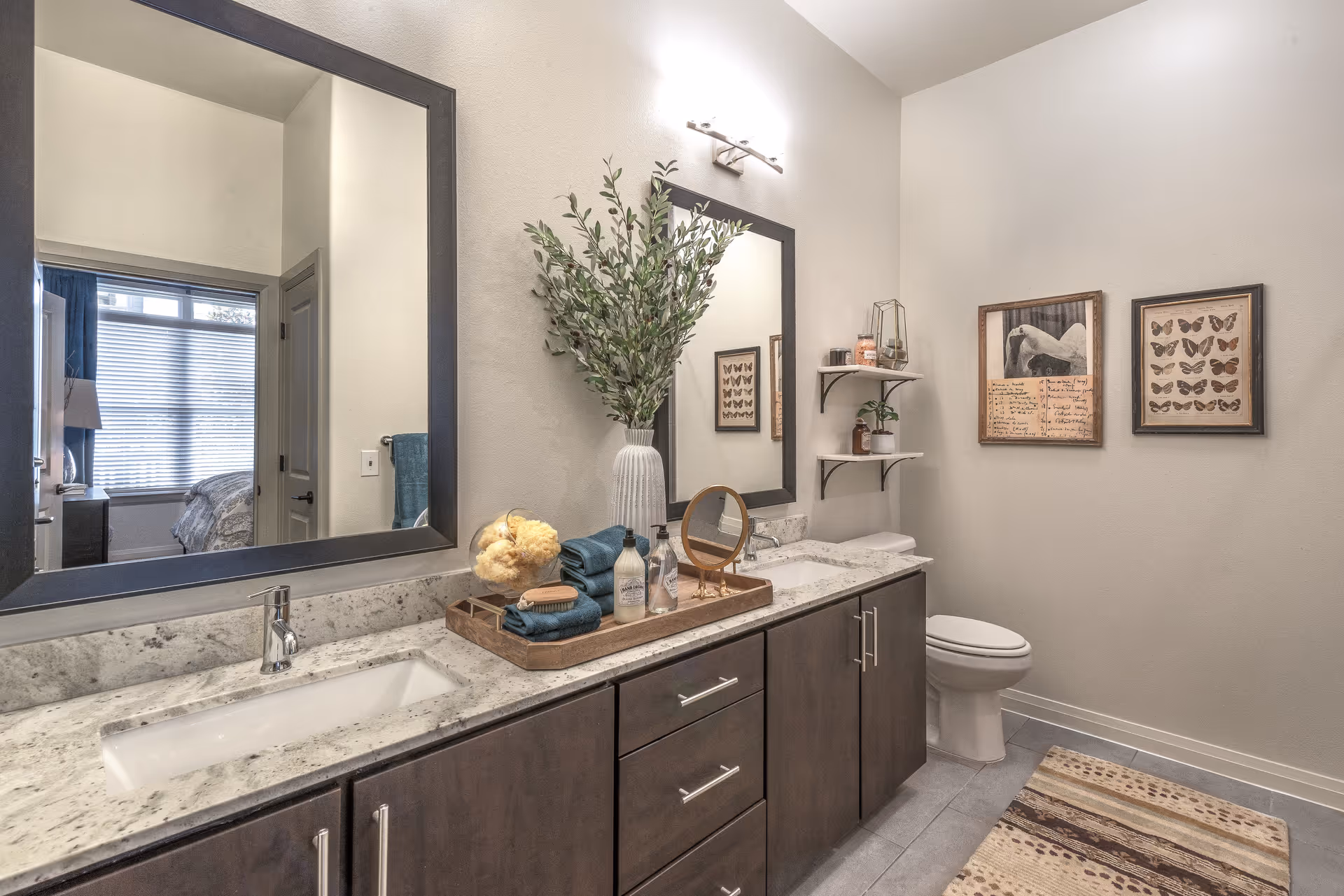 A modern bathroom with a double sink vanity featuring a marble countertop and dark wood cabinets. Above the sinks are two large mirrors with a light fixture above one. On the countertop is a wooden tray holding towels, soap dispensers, a sponge, and a small mirror. A tall vase with green leafy branches is placed between the sinks. To the right, there is a toilet and two framed butterfly and nature-themed artworks on the wall. Two small shelves hold decorative items and a plant. A patterned rug is on the tiled floor. A doorway reflects a bedroom with a window and bed visible.