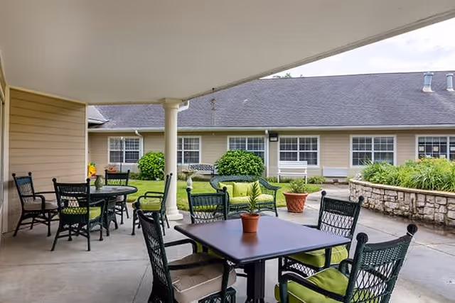 Covered outdoor patio area with black metal tables and chairs featuring green cushions. There are potted plants on the tables and a stone planter with greenery in the background. The patio overlooks a courtyard with bushes and a beige building with multiple windows.