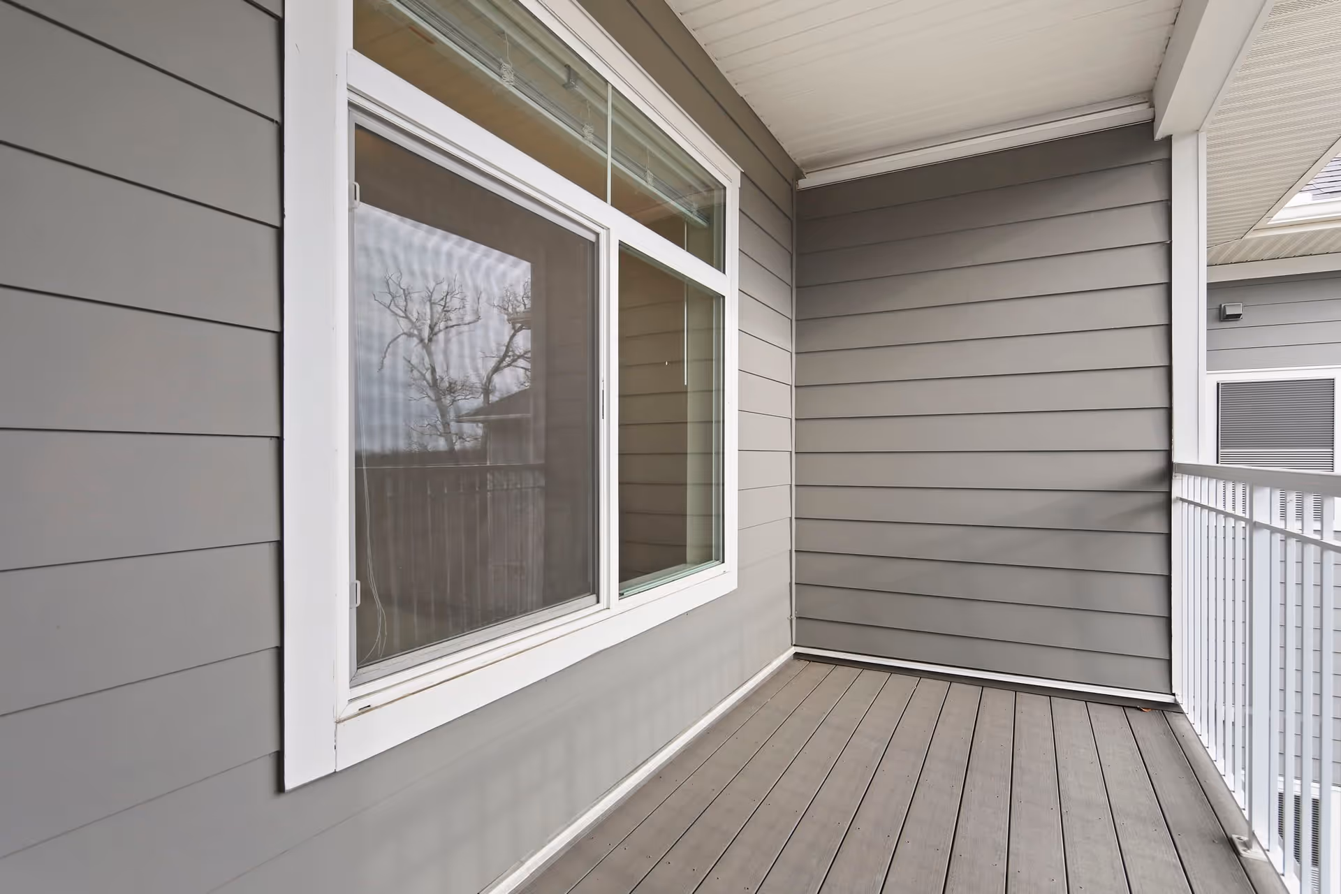 A small outdoor balcony area with gray wooden siding walls, a white-framed window, and a white metal railing. The floor is made of gray wooden planks and the balcony is covered by a white ceiling.
