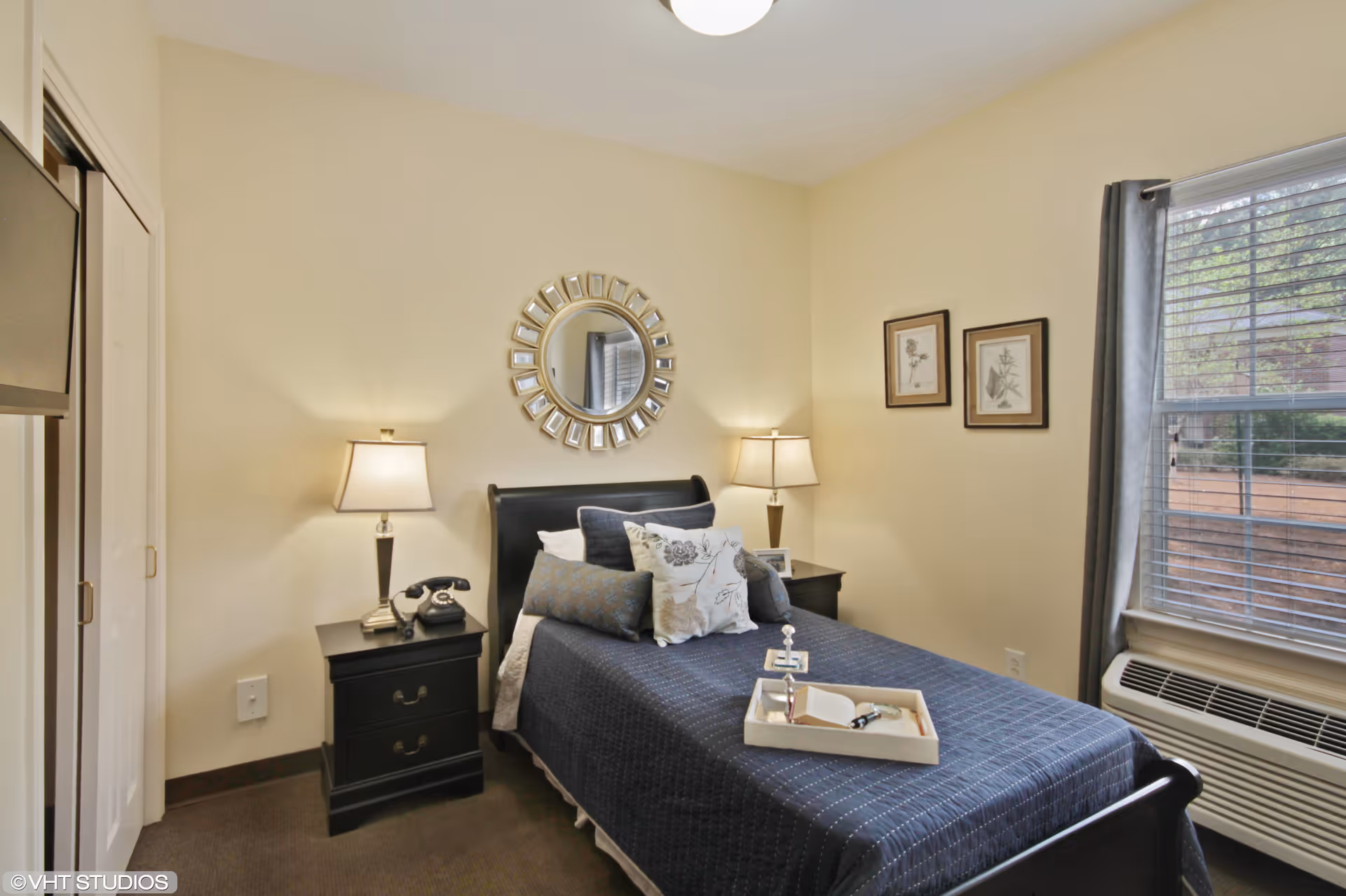 A cozy bedroom with a single bed covered in a dark blue quilt and multiple pillows. There are two black nightstands on either side of the bed, each with a lamp. A round decorative mirror hangs above the bed, and two framed pictures are on the adjacent wall. A window with gray curtains lets in natural light, and an air conditioning unit is installed below the window. A tray with a book and a small decorative item is placed on the bed.