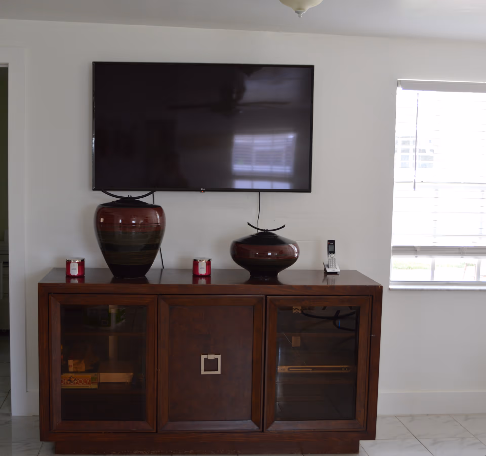 A wall-mounted flat screen TV above a wooden cabinet with glass doors. On top of the cabinet are two decorative vases, two small red candles, and a cordless phone. A window with blinds is on the right side, letting in natural light.