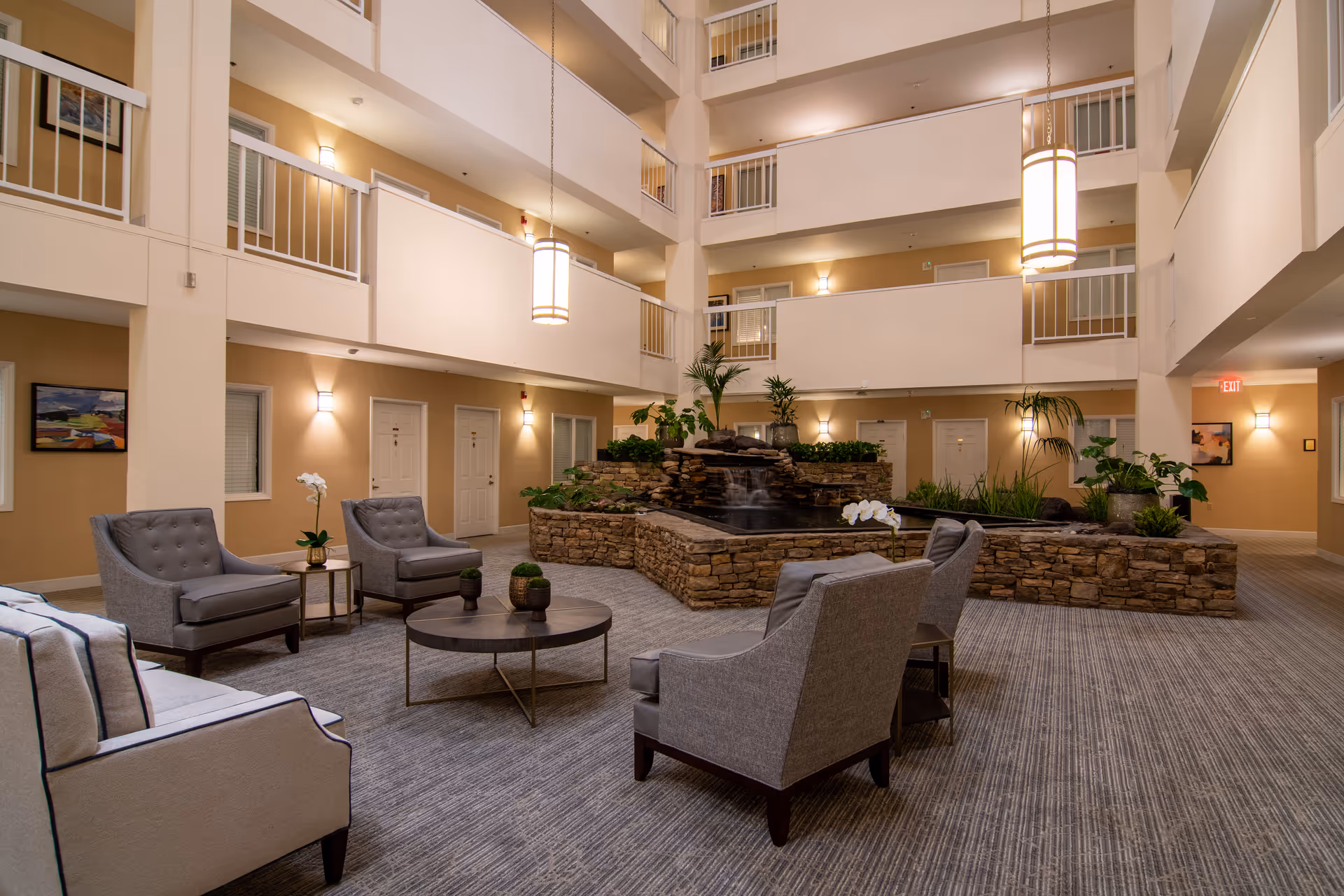 Indoor common area of a retirement community featuring comfortable seating with armchairs and a sofa arranged around a round coffee table. In the center, there is a decorative stone water feature with plants. The space is well-lit with hanging pendant lights and wall sconces, and multiple doors and balconies are visible on the upper floors.