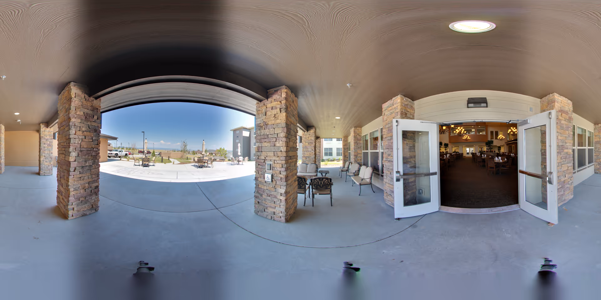 Covered outdoor patio area with stone pillars and seating, leading to an open courtyard with tables and chairs under a clear blue sky. Double glass doors open into an interior dining area with tables and chandeliers visible inside.