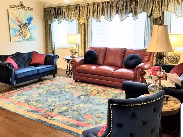 A cozy living room with a red leather sofa, two dark blue upholstered chairs, and a matching loveseat. The seating surrounds a colorful area rug with floral patterns. There are two side tables with decorative lamps and a vase of flowers. A large window with patterned curtains lets in natural light, and a colorful abstract painting hangs on the wall.