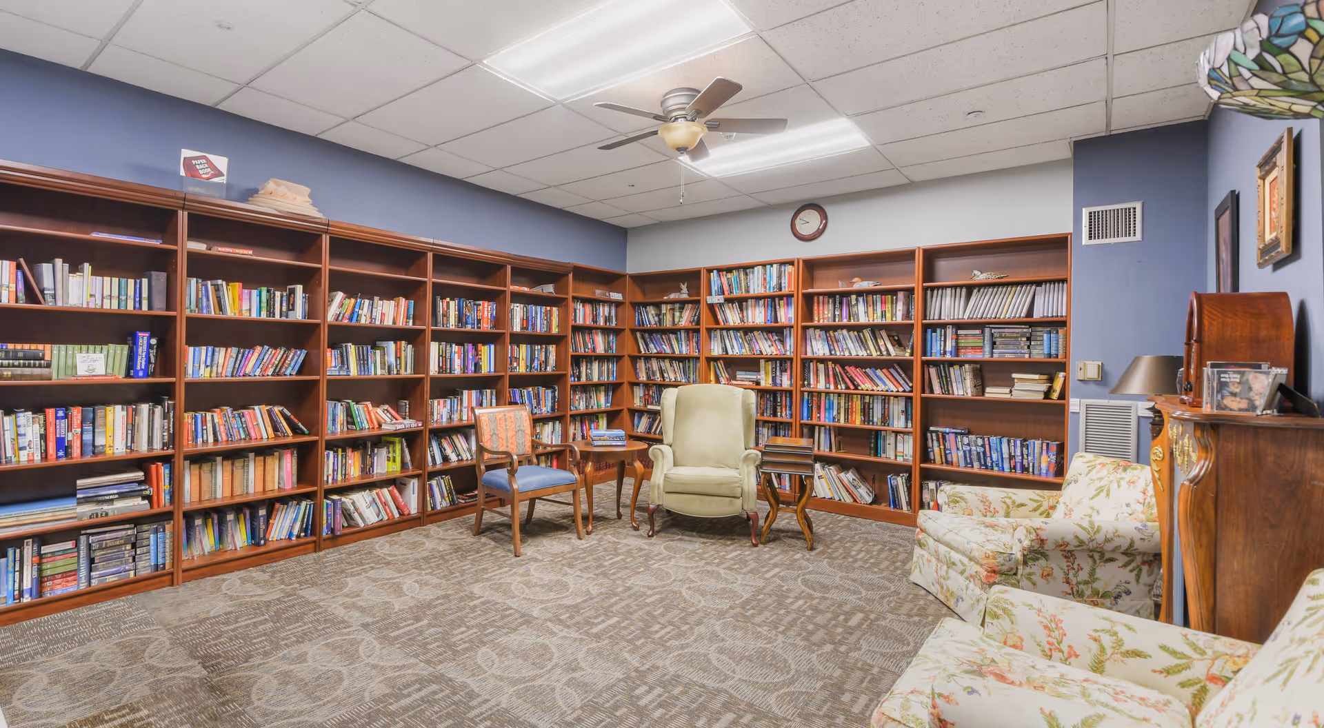 A cozy library room in a senior living community with wooden bookshelves filled with books lining two walls. The room has a ceiling fan with lights, a clock on the wall, and several comfortable chairs including a green armchair, a wooden chair with cushions, and floral upholstered armchairs. There are small wooden side tables placed near the chairs and a vintage-style wooden cabinet with a lamp and framed photos on top.