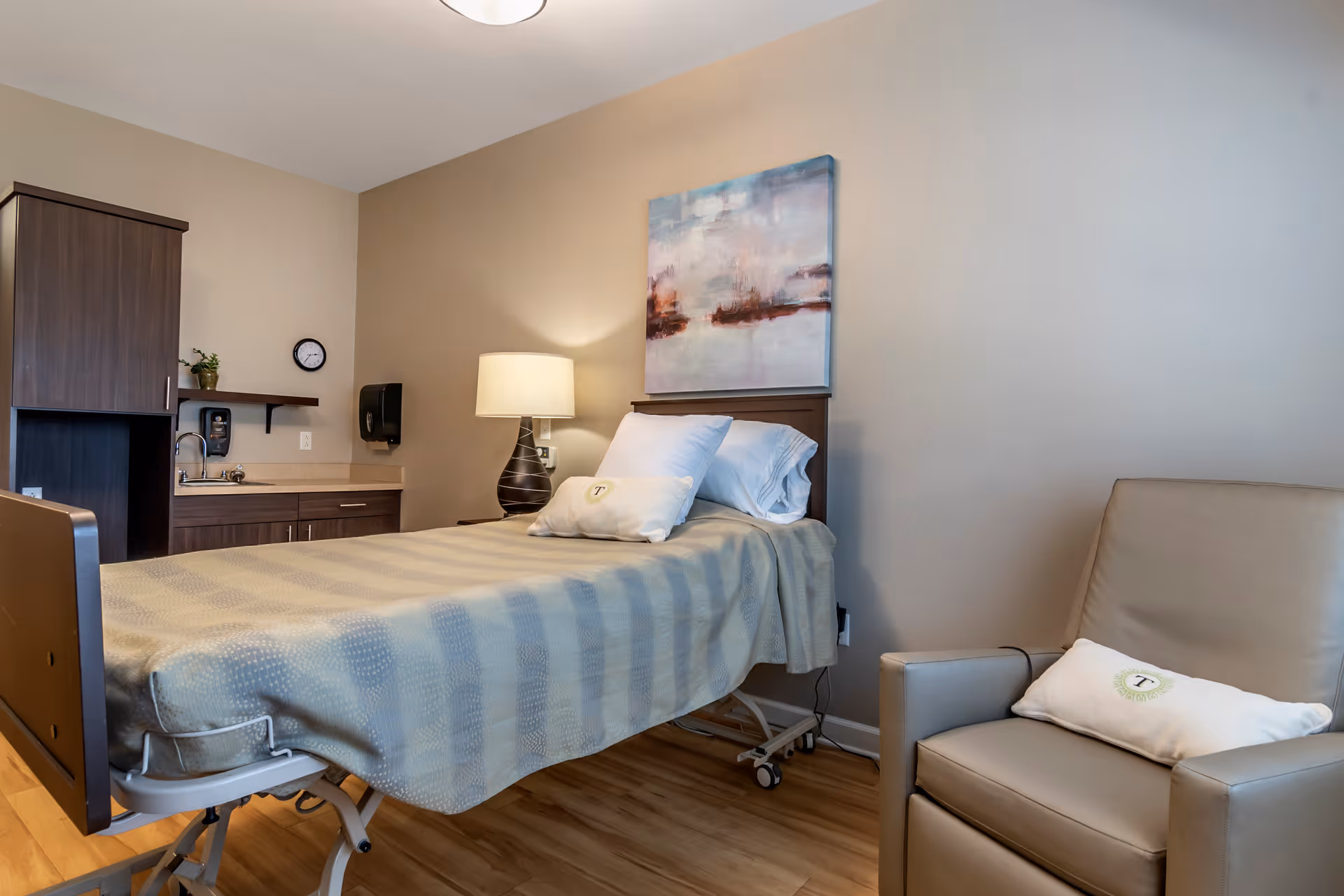 A clean and well-lit room with a single hospital-style bed covered with a beige and light blue patterned blanket. The bed has white pillows, one with a logo. Next to the bed is a wooden nightstand with a modern lamp and a large abstract painting on the wall above the bed. To the left, there is a small sink area with dark wood cabinets, a clock, and a soap dispenser. On the right side of the image, there is a beige armchair with a white pillow featuring a logo. The floor is wooden, and the walls are painted in neutral tones.