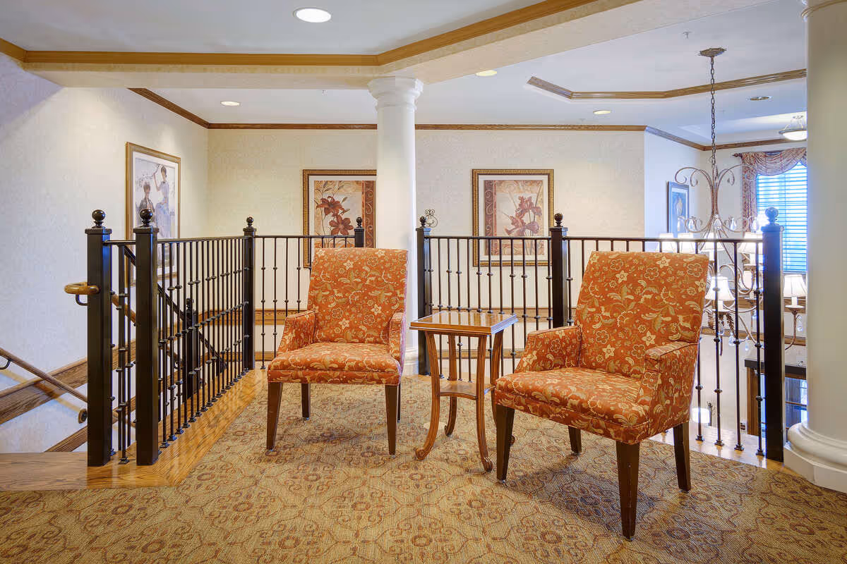 A cozy seating area with two orange patterned armchairs and a small wooden side table between them. The area is carpeted with a patterned rug and is bordered by a black metal railing. The background features framed floral artwork on cream-colored walls, a white column, and a chandelier hanging from the ceiling.