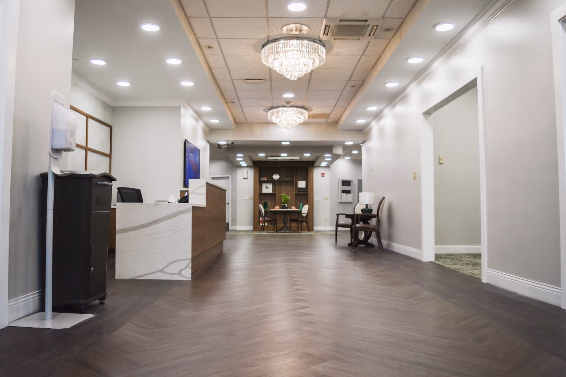 A spacious, well-lit reception area with a modern front desk on the left, two chandeliers hanging from the ceiling, a small table with chairs and decorative plants at the far end, and a chair with a lamp on a side table along the right wall.