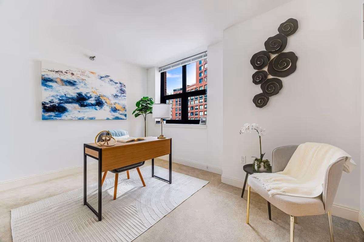 A bright, modern office space with a wooden desk and gray chair on a textured white rug. A large abstract blue and white painting hangs on the wall to the left. A window shows a view of a red brick building outside. To the right, there is a light gray armchair with a white throw blanket and a small round side table with a white orchid plant. Decorative black spiral wall art is mounted above the armchair.