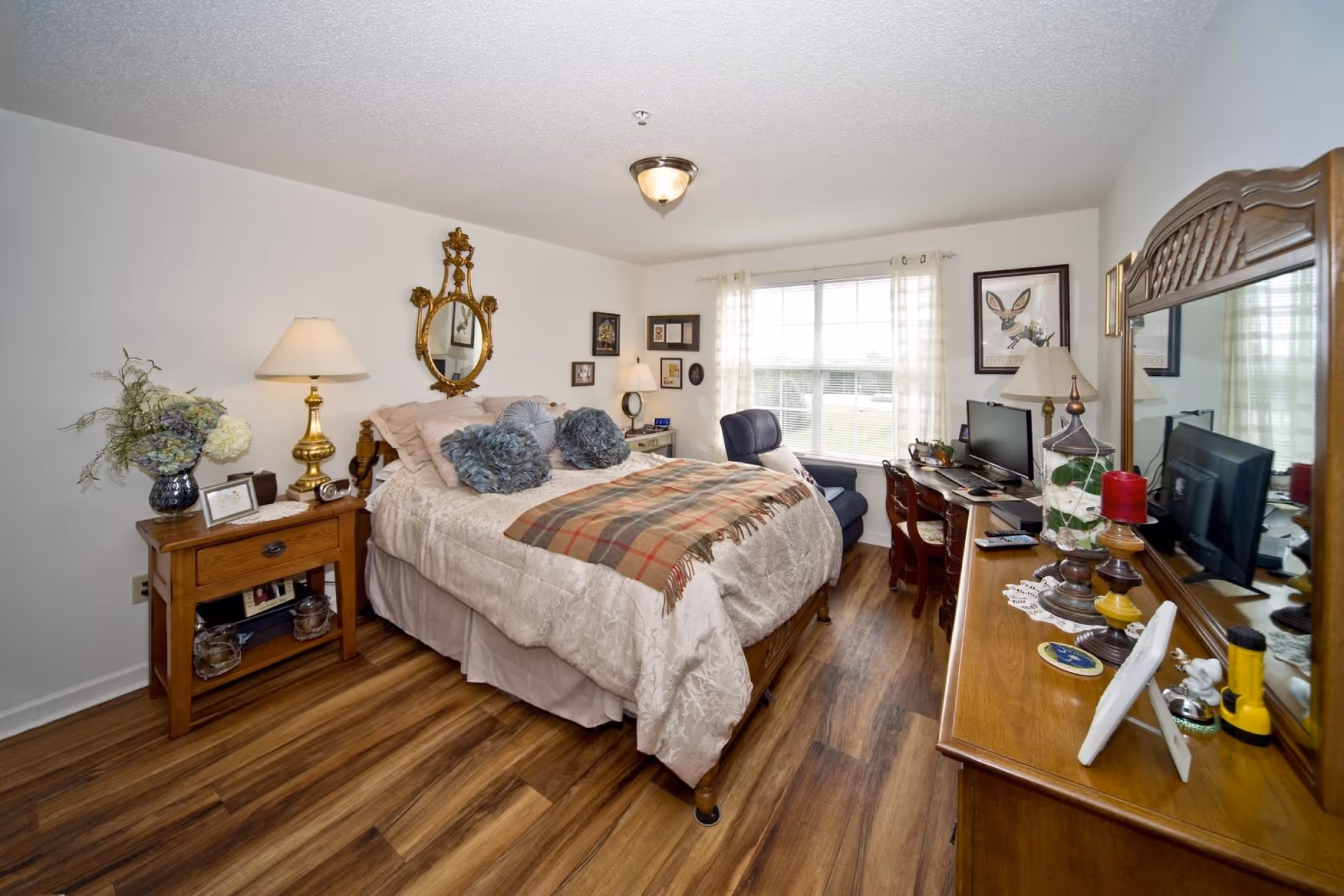 A cozy bedroom with a large bed covered in beige bedding and decorative pillows. There is a wooden nightstand with a lamp and flowers on the left side of the bed. A gold-framed oval mirror hangs above the bed. A window with sheer curtains lets in natural light. On the right side, there is a wooden dresser with a mirror, a TV, and various decorative items. A blue armchair and a wooden desk with a chair are positioned near the window. The room has wood flooring and white walls.