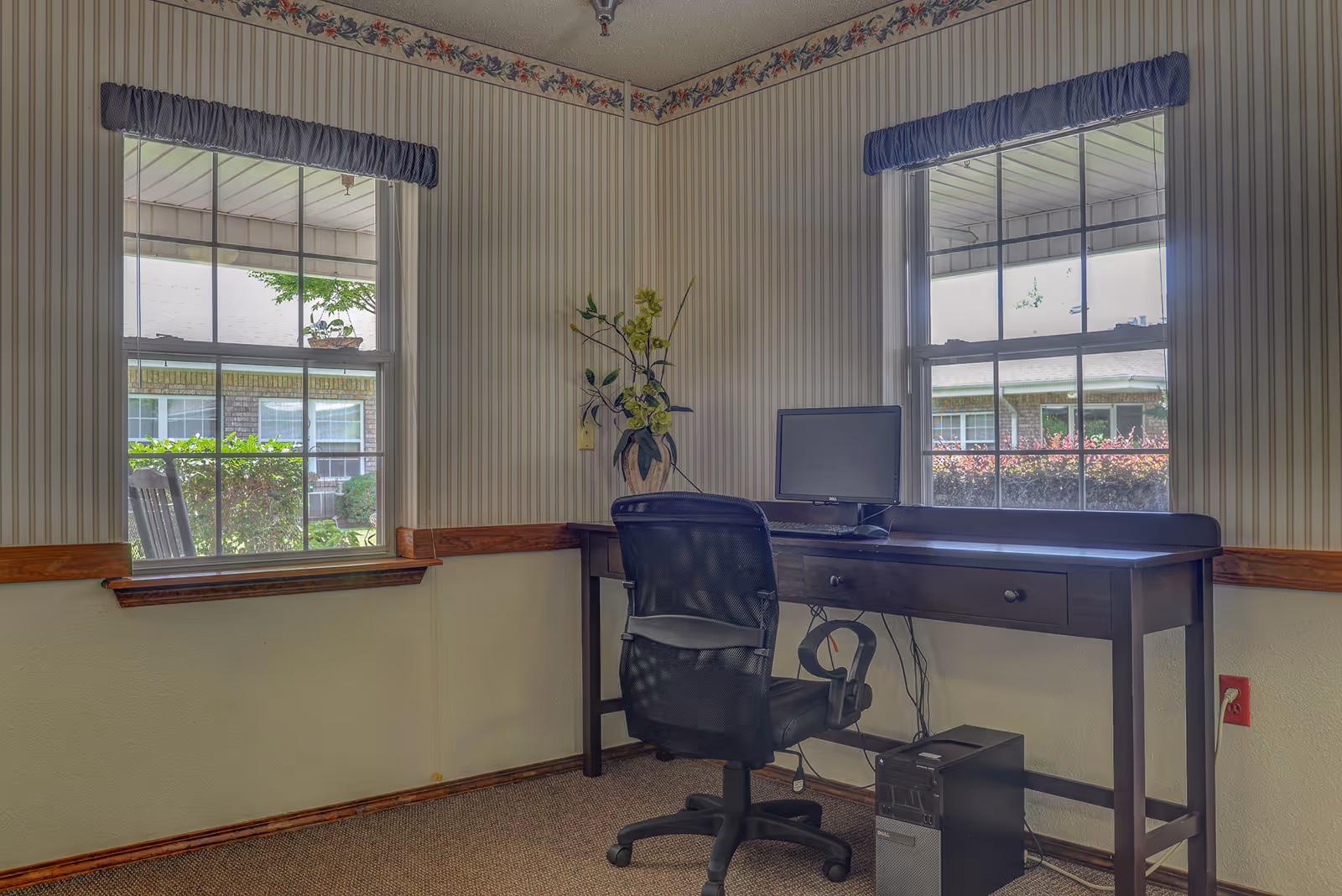 A small office area with a dark wooden desk and a black mesh office chair. On the desk is a computer monitor and keyboard, with a desktop tower placed on the floor underneath. Two windows with blue valances provide natural light and a view of greenery and a brick building outside. The walls have striped wallpaper with a floral border near the ceiling.