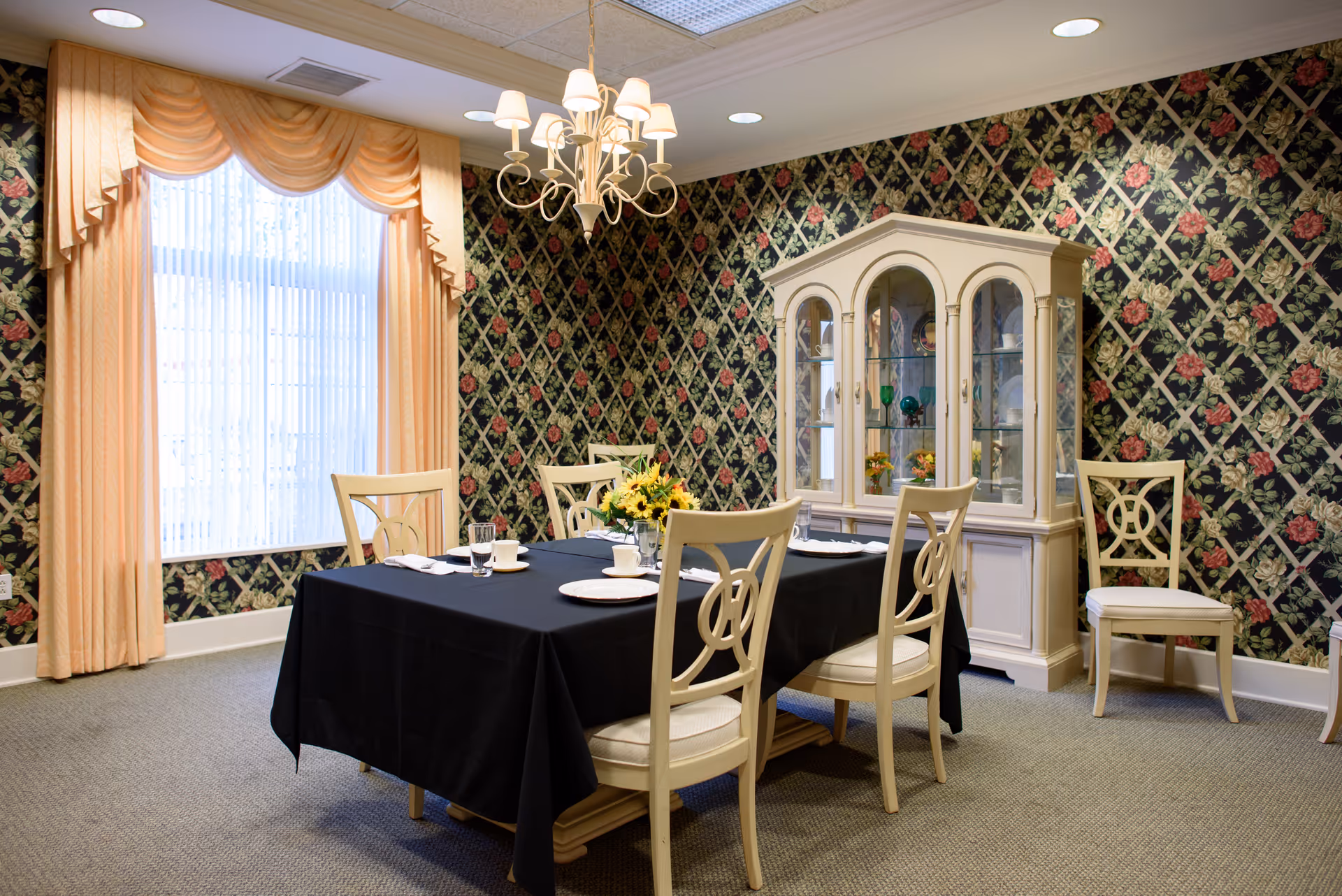 A dining room with a rectangular table covered by a black tablecloth, set with white plates, cups, and glasses. There are six cream-colored chairs around the table. The room features floral patterned wallpaper with a lattice design, a large window with peach-colored curtains, a chandelier hanging from the ceiling, and a cream-colored china cabinet displaying decorative items.