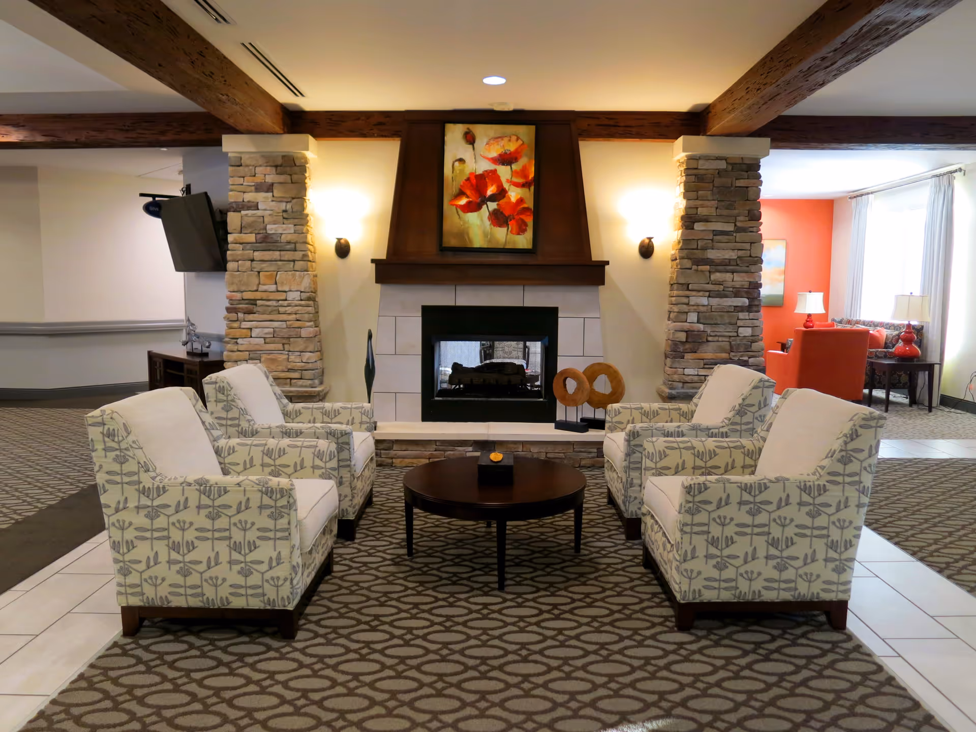 A cozy assisted-living common room with four patterned armchairs arranged around a coffee table facing a fireplace beneath a floral painting.