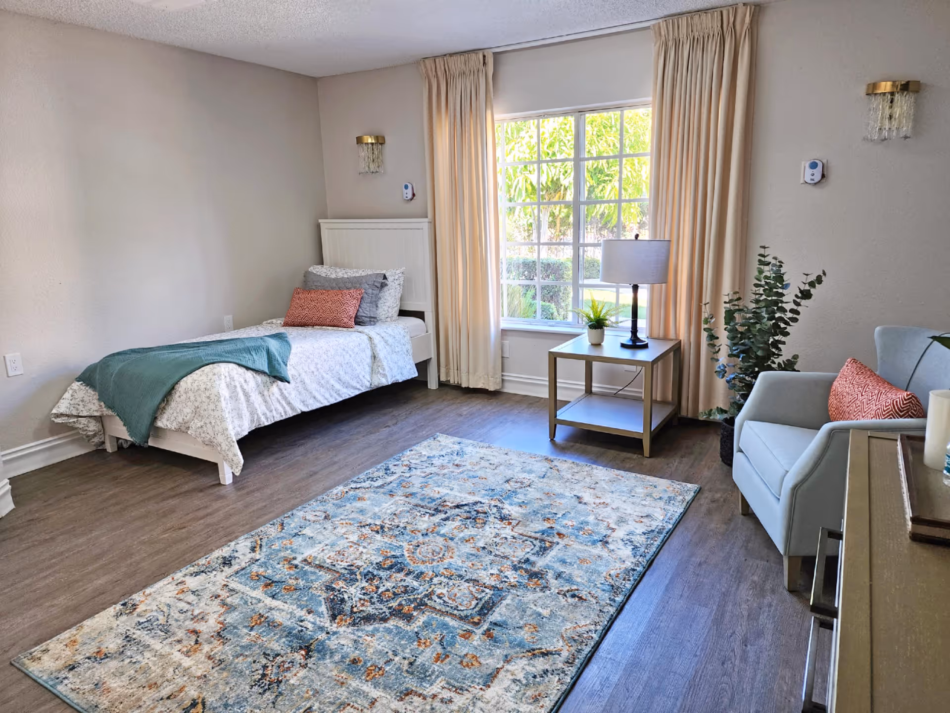 A bright and cozy bedroom with a single bed against the left wall, covered with a white patterned bedspread, a teal throw blanket, and a coral accent pillow. Next to the bed is a window with cream-colored curtains letting in natural light. A wooden side table with a lamp and a small plant sits in front of the window. To the right, there is a light blue armchair with a coral pillow and a tall green plant beside it. The floor is wood with a large patterned area rug in shades of blue, cream, and orange.