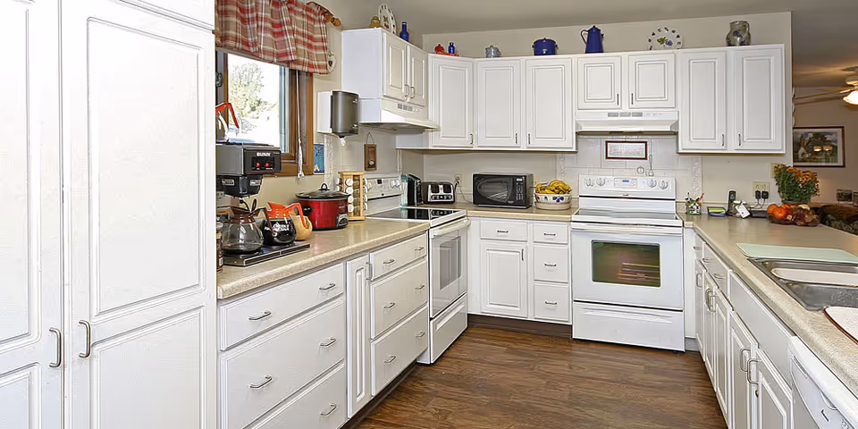 A bright kitchen with white cabinets and drawers, a white stove and oven, a microwave, a coffee maker, and various kitchen items on the countertops. There is a window with a red plaid valance above the counter, and the floor is a dark wood laminate.