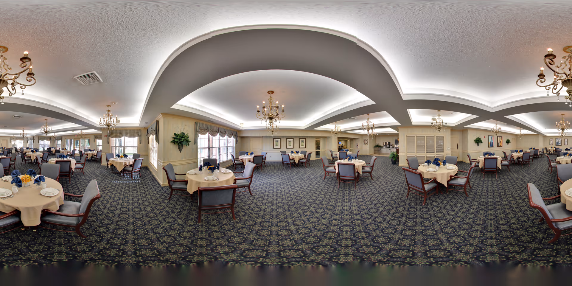 A spacious and elegant dining room in Oakmont Parkway Senior Community with round tables covered in beige tablecloths, each set with plates, napkins, and floral centerpieces. The room features patterned carpet, large windows with valances, chandeliers hanging from a ceiling with recessed lighting, and comfortable upholstered chairs arranged around the tables.
