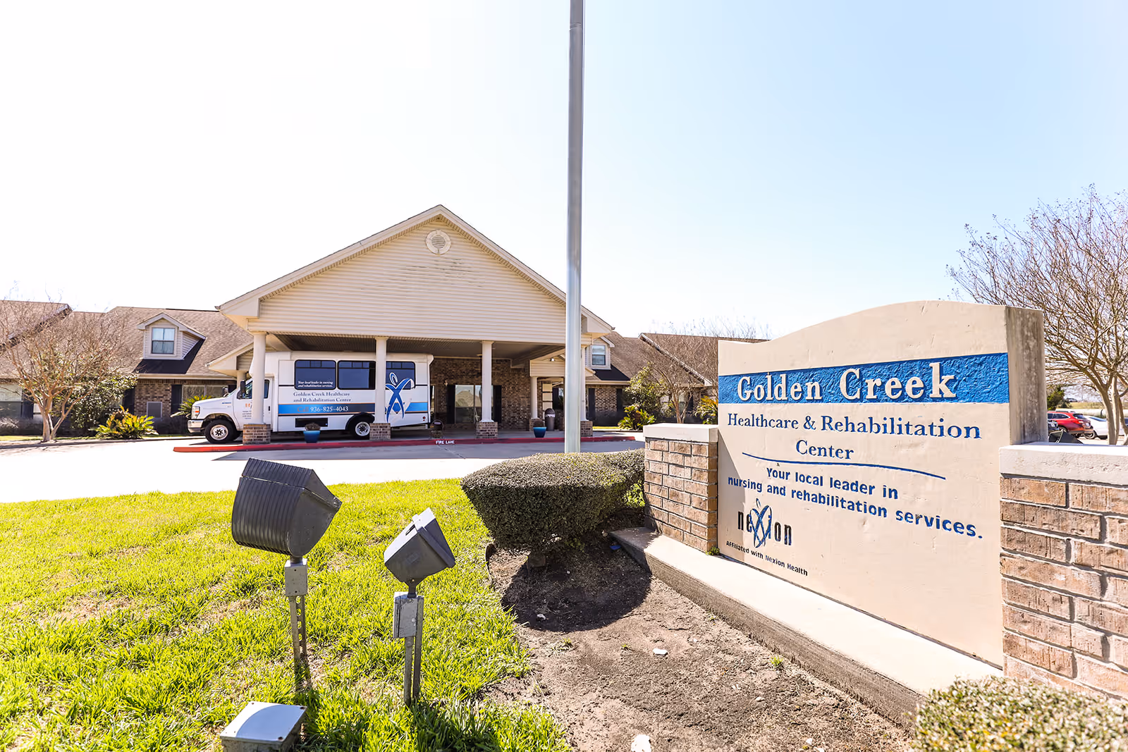 Exterior view of Golden Creek Healthcare and Rehabilitation Center building with a sign in the foreground displaying the facility's name and slogan. A shuttle van is parked under the covered entrance, and there are some trees and landscaping around the building.