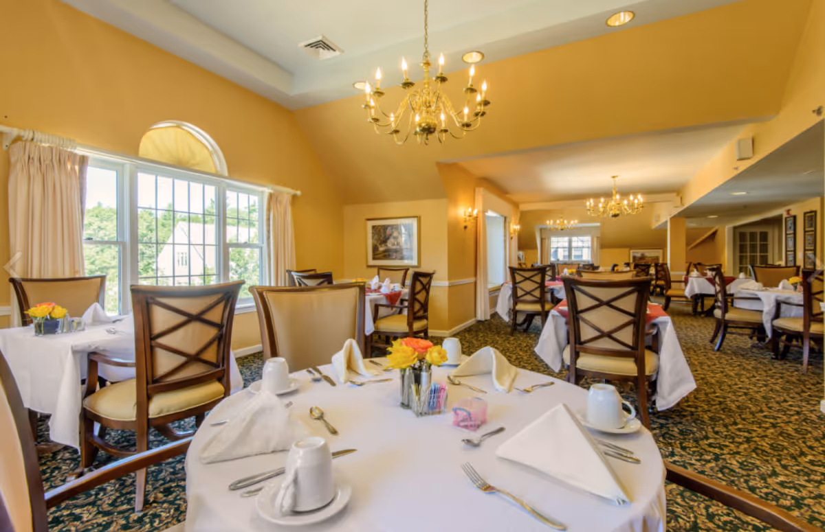 A bright and elegant dining room with multiple round tables covered with white tablecloths, set with folded napkins, cups, and silverware. The room features large windows with cream-colored curtains, patterned carpet, and gold chandeliers hanging from the ceiling. There are wooden chairs with cushioned seats around the tables.
