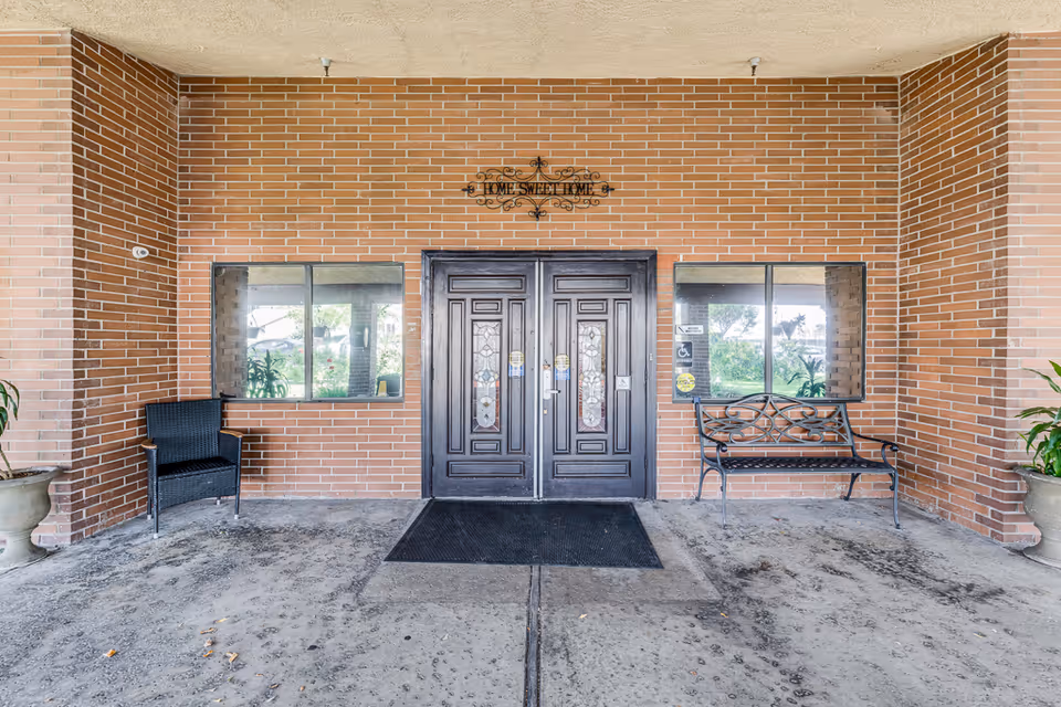 Entrance to a building with double dark wooden doors featuring decorative glass panels. The walls are made of reddish-brown bricks. Above the doors is a decorative metal sign that reads 'Home Sweet Home'. There are two windows on either side of the doors, a black wicker chair to the left, and a black metal bench to the right. Two large potted plants flank the entrance area.