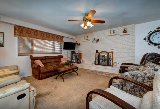 A cozy living room with a brown sofa, floral armchair, beige recliner, and a white cushioned chair. There is a wooden coffee table with a plant on it in the center. The room features a white brick fireplace with decorative items on the mantel and a screen in front. A ceiling fan with lights is mounted on the ceiling. A flat-screen TV is mounted on the wall near a bookshelf filled with books. The window has wooden blinds and a patterned valance.