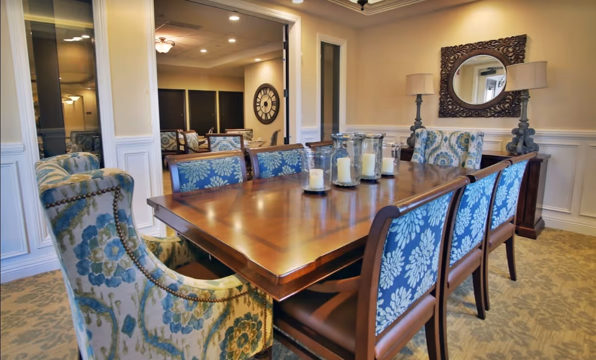 A well-lit dining room with a polished wooden table surrounded by eight upholstered chairs featuring blue floral patterns. On the table, there are several glass candle holders with white candles. The room has beige walls with white wainscoting, a decorative mirror with an ornate frame, two matching lamps on a sideboard, and a large clock visible in an adjacent room.