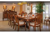 A well-lit dining room with a large wooden dining table set for a meal, surrounded by eight wooden chairs with cushioned seats. The table is decorated with a floral centerpiece and place settings including plates, cups, and utensils. The room features beige walls, a sideboard with decorative items, framed pictures on the walls, a large potted plant near a window with white blinds, and two wall-mounted lantern-style lights.