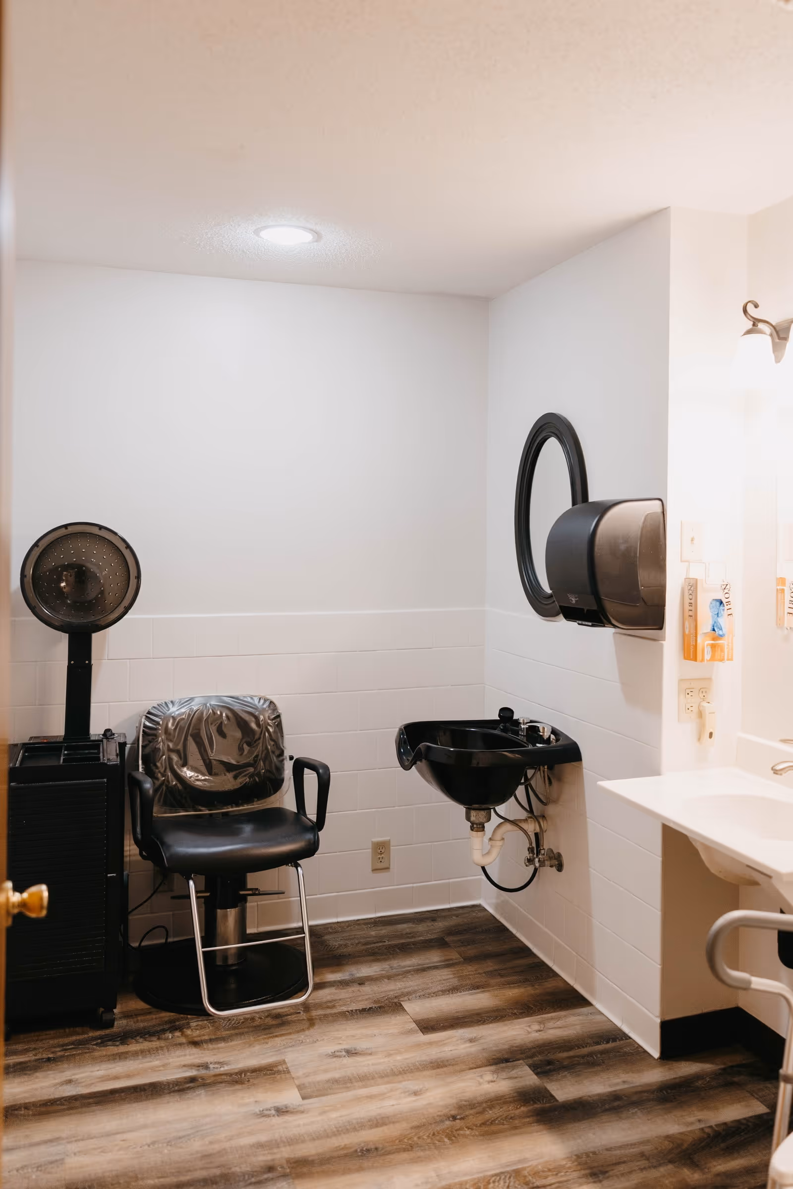 Interior of a small salon or grooming room with a black salon chair covered in plastic, a black hair dryer hood, a black sink mounted on the wall, a round mirror, a soap dispenser, and a white countertop with a sink. The floor is wood-patterned and the walls are white with white tiles halfway up.