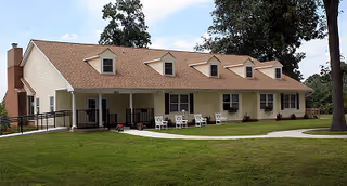 Single-story residential building with beige siding and a brown shingled roof, featuring multiple dormer windows and a chimney. The building has a covered porch with several white rocking chairs. It is surrounded by a well-maintained green lawn and trees under a partly cloudy sky.