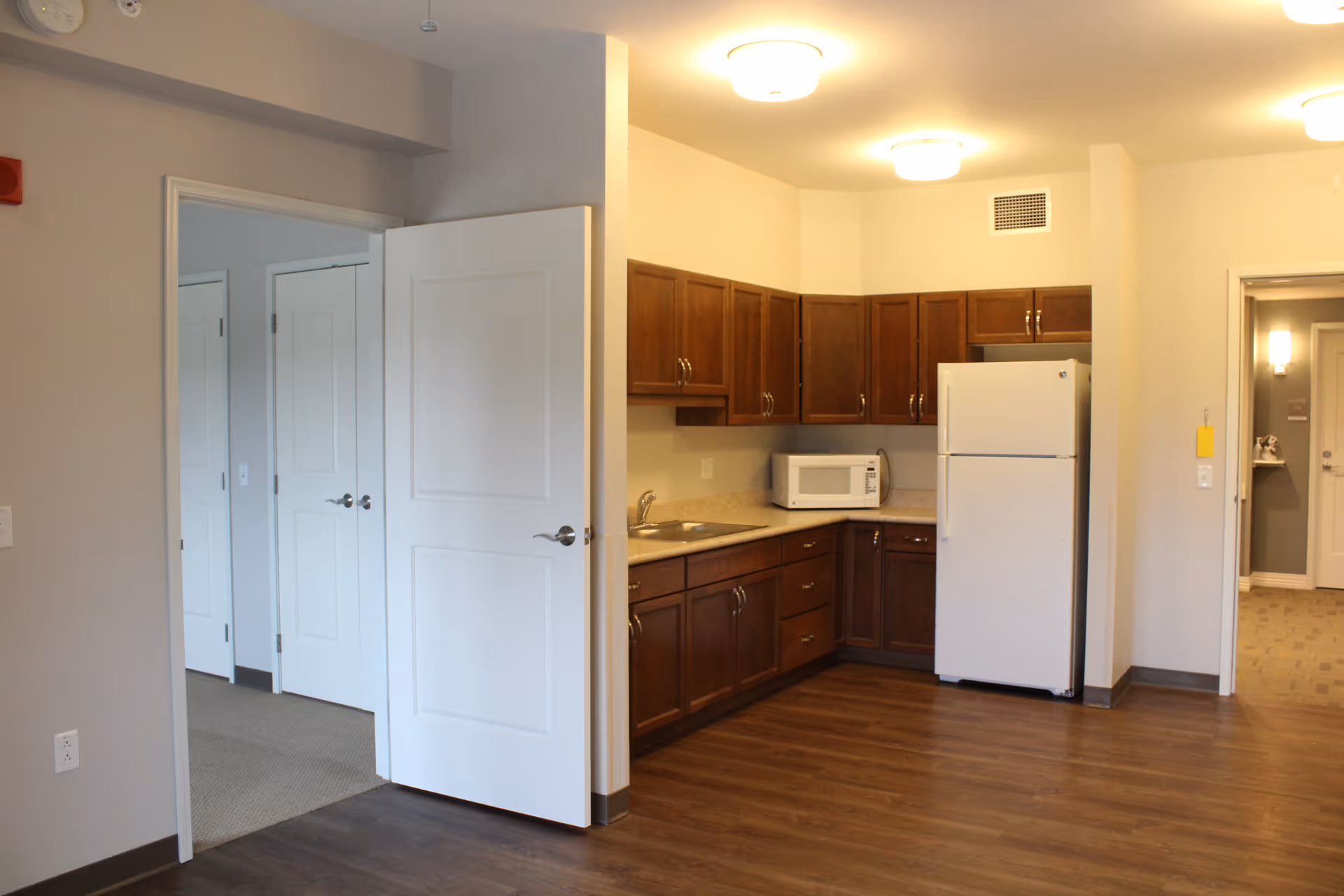 Interior view of a senior living facility kitchen area with wooden cabinets, a white refrigerator, a microwave on the counter, and a sink. The kitchen has wood flooring and is adjacent to a hallway with open white doors leading to other rooms.