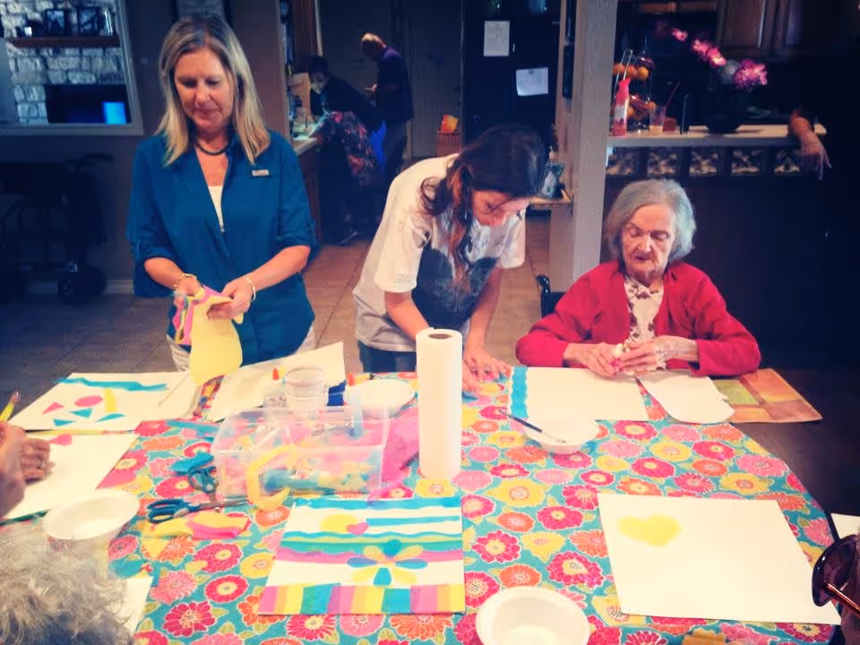 A group of people engaged in an arts and crafts activity at a table covered with a colorful floral tablecloth. An elderly woman in a red cardigan is seated and working on a project, while two other women stand nearby assisting or participating. Various craft supplies like paper, scissors, and glue are spread out on the table. The setting appears to be a communal indoor area in a senior living facility.