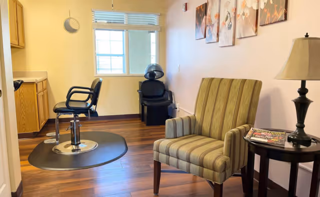 Interior salon area with two styling chairs and a hooded hair dryer, plus a striped armchair beside a side table with a lamp and magazines.