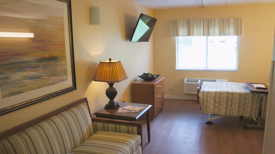 A well-lit room with a single bed covered in a striped quilt, a wooden dresser with a decorative bowl, a wall-mounted TV, a window with blinds and a striped valance, a side table with a lamp and magazines, and a striped couch beneath a large framed abstract painting.