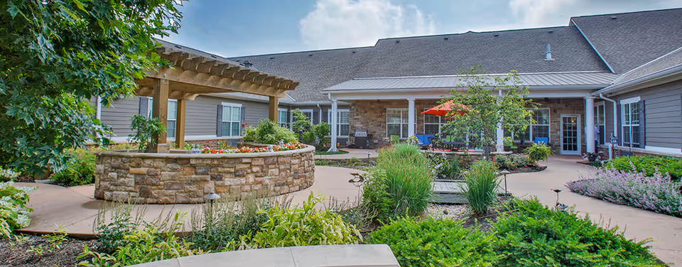 Outdoor courtyard area of a senior living facility with a stone planter, wooden pergola, various plants and shrubs, and a building with a covered patio and seating area in the background under a partly cloudy sky.