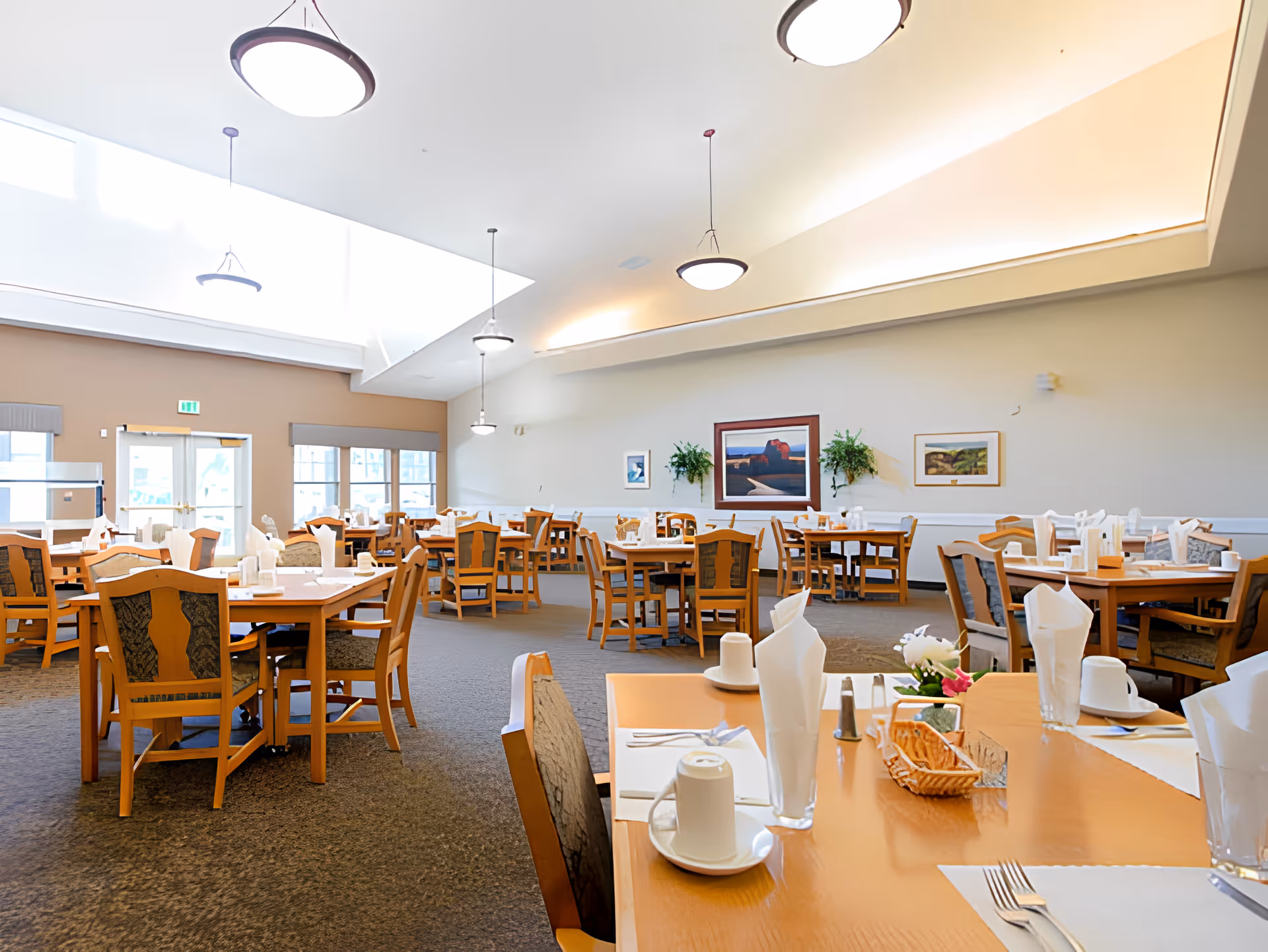 A bright and spacious dining room with multiple wooden tables and chairs arranged neatly. Each table is set with white napkins, cups, and cutlery. The room has high ceilings with modern hanging light fixtures and large windows allowing natural light to fill the space. There are framed pictures and plants on the walls, creating a welcoming atmosphere.