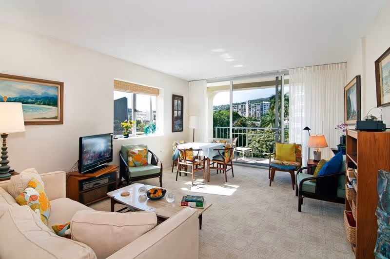 Sunlit living room with a sofa, armchairs, coffee table, TV and a small dining table by sliding glass doors opening onto a balcony and greenery.
