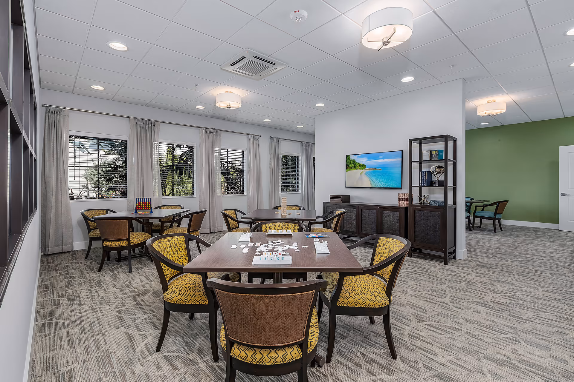 A bright and spacious common area with several tables and chairs arranged for games and social activities. One table has a game of Rummikub set up, and another table has a Connect Four game. The room features large windows with white curtains, a flat-screen TV mounted on the wall displaying a beach scene, and a shelving unit with decorative items. The walls are painted white and green, and the floor is carpeted with a patterned design.
