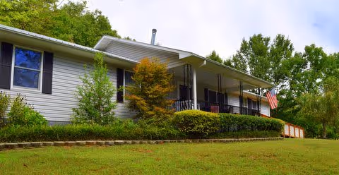 Single-story residential building with a covered porch, surrounded by green shrubs and trees, with an American flag displayed near the entrance under a partly cloudy sky.