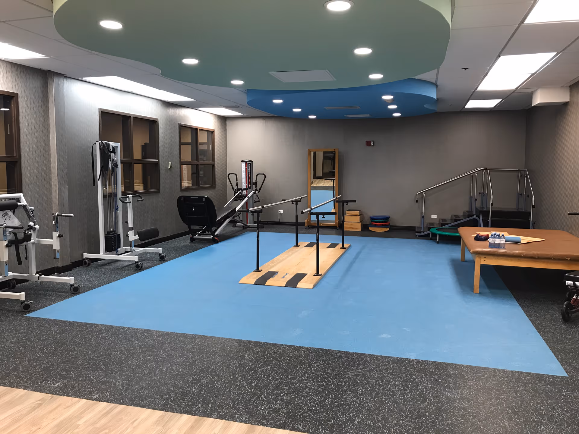 Interior therapy/fitness room with parallel walking bars, exercise equipment, and a treatment table on blue flooring.